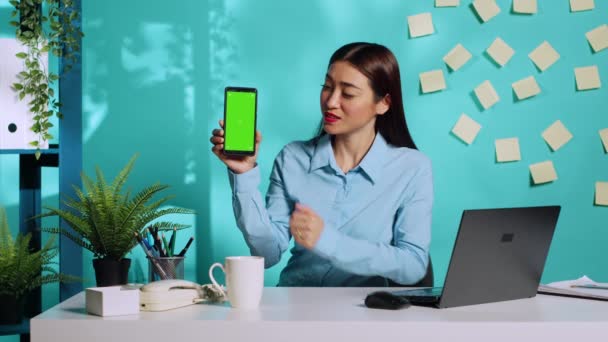 クロマキーグリーンの画面でスマートフォンを持っている陽気なアジアの事務員の女性は 情報を提示します 青のスタジオを背景にカラフルなリラックスした職場で専門秘書 — ストック動画