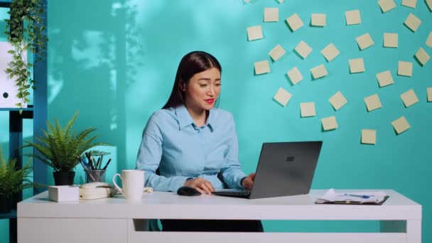 同僚とのエキサイティングな会話をリラックスしているオンラインビデオコールの陽気なオフィスワーカー 笑顔の従業員が青いスタジオの背景の上に現代の職場で机に座って — ストック動画