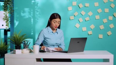 İnternetteki video konferans toplantısında müdürden gelen bilgiler üzerine Asyalı sekreter pano evraklarını tamamlıyor. Modern ofisteki çalışan kadın mavi stüdyo geçmişinden üstün.