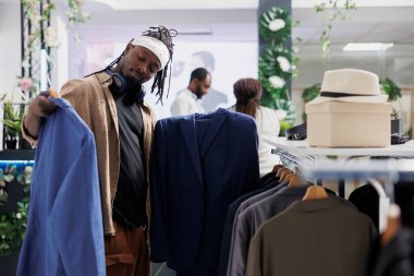 Alıcı iki ceket arasında karar verirken giysi mağazasında resmi kıyafet seçiyor. Alışveriş merkezinde kumaş ve tarzı incelerken askılarda ceketler tutan Afrikalı Amerikalı bir adam.