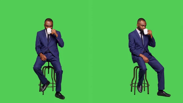 椅子でコーヒーを提供するスタートアップ起業家は フルボディグリーンの背景を持つスタジオに座っています 男性オフィス労働者は カメラでカフェインのリフレッシュを飲んで 飲料を飲む — ストック写真