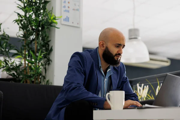アラブのマーケティングマネージャーは ラップトップ上の販売戦略でビジネスオフィスを起動します 同僚のスペースでソファに座っている間 男起業家はプロジェクトレポートを分析します — ストック写真