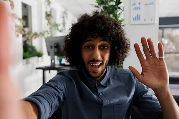 Start up company employee recording video for customer on smartphone front camera and waving hi. Smiling arab entrepreneur greeting coworker while communicating online on mobile phone