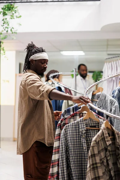 アフリカ系アメリカ人の男性が洋服店で流行の服を探している ショッピングモールのファッションブティックでカジュアルなアパレルを選択するためにラックにぶら下がっているシャツを通して顧客の閲覧 — ストック写真