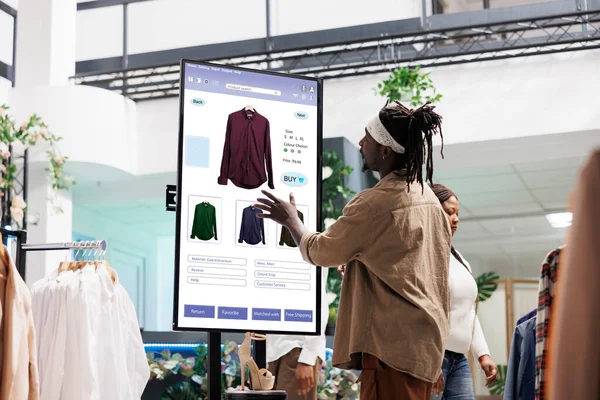 Customer using touch display shopping for fashion items in clothing store, interactive board. African american man choosing stylish clothes with different designs, modern kiosk service.