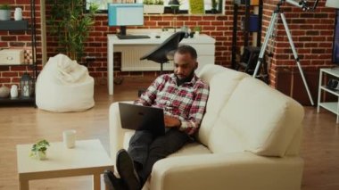 Evde çalışırken laptoptan veri girişi yapan rahat bir serbest çalışan. Afrikalı Amerikalı tele-işçi kanepede uzanıp şık, sıcak bir apartman dairesinde uzaktan kumandayla iş yapıyor.