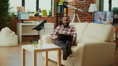 Afrikalı Amerikalı yönetici uzaktan çalışan iş arkadaşlarıyla çevrimiçi video konferansı düzenliyor. Modaya uygun bir dairede koltukta uzanmış televizyon yönetmeniyle iş arkadaşlarıyla görüntülü konuşma.