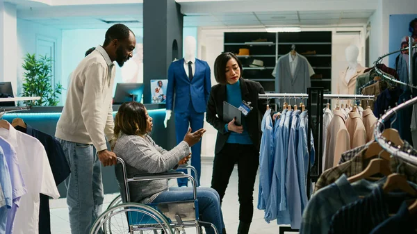 車椅子ショッピングモールでアフリカ系アメリカ人女性 衣料品店で現代の服コレクションを検討 身体障害のある男性買い物客カジュアルウェア ショッピングモールを購入 — ストック写真