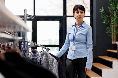 Süslü kıyafetlerle çalışan bir mağaza çalışanı, alışveriş merkezindeki askılara şık ürünler koyuyor. Modern butikte günlük kıyafet düzenleyen şık gömlekli neşeli menajer.