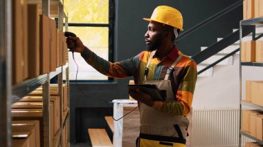 Afro-Amerikalı adam depoda barkodları kontrol ediyor, tarayıcı ve tabletle lojistik üzerinde çalışıyor. Raflardaki kutular ile ürün kalite denetimi gözden geçiriliyor, ikmal denetimi yapılıyor.