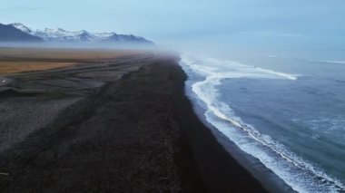 İzlanda siyah kumsalı drone çekimi, Atlantik kıyılarına dalgaların çarptığı güzel sahil şeridi. Karlı dağları ve kumsalda siyah kumları olan kuzey manzarası, manzaralı bir yol. Yavaş çekim.