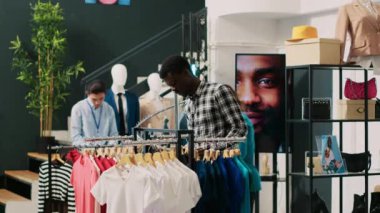 Afro-amerikan alışverişçi, temel gömleği kontrol ediyor, giyim mağazasındaki kumaş ürünlerini analiz ediyor. Modern butikte alışveriş manyağı bir müşteri şık kıyafetler alıyor. Moda konsepti