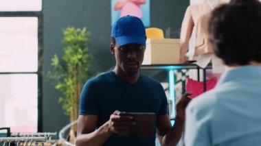 Kısa saç teslimatı raporunu imzalayan yönetici giyim mağazasında Afro-Amerikan kuryeyle lojistiği tartışıyor. Modern butikte nakliye için paketler hazırlayan işçiler. Moda konsepti