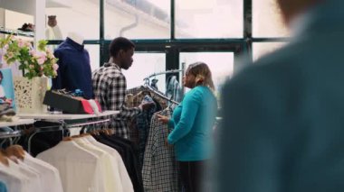 Afro-Amerikan çift şık ürünlere bakıyor, giyim mağazasındaki kumaşlara bakıyor. Gülümseyen müşteriler alışveriş yapmaktan hoşlanıyor, modern butikten moda kıyafetler ve aksesuarlar alıyorlar.