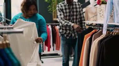 Yeni moda koleksiyonunu kontrol eden Afrikalı Amerikalı müşteriler modern butikte günlük kıyafet alışverişi yapıyorlar. Neşeli çift kıyafet analizi yapıyor, şık ürünler alıyor. Moda konsepti