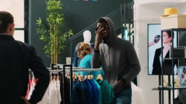 Afro-Amerikan hırsızı elbise mağazasında güvenlik görevlisi tarafından yakalanarak moda kıyafetleri çalmaya çalışıyor. Asyalı koruma soyguncunun modaya uygun malları iade etmesini yoksa polisi arayacağını söyledi.