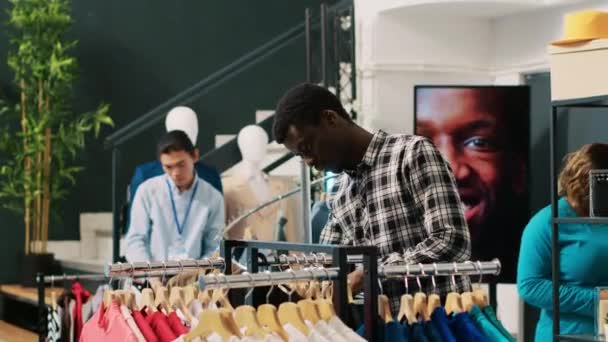 时尚客户购买时尚服饰 分析服装店的商品材料 黑人男子看着衣架与新的时尚系列 购买基本的服装 — 图库视频影像