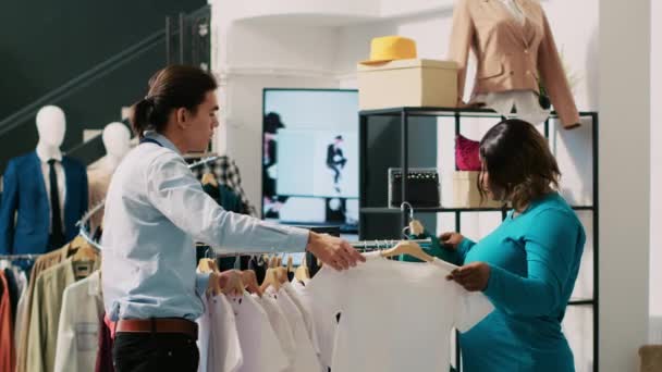 非洲裔美国人顾客在现代精品店寻求工人的帮助 购买时尚商品 亚洲员工在服装店安排衣架和时髦服装 时尚概念 — 图库视频影像