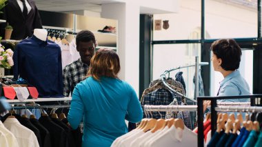 Afrika kökenli Amerikalı çift şık gömleklere bakıyor, modern butikte moda ürünlerle dolu askıları kontrol ediyorlar. Neşeli müşteriler giyim mağazasından almadan önce kıyafet malzemelerini kontrol ediyorlar.