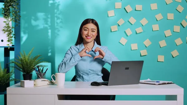 手のハート型のサインを示す幸せなチッパー暖かいアジアのビジネス女性 オフィス書記を保持アップ愛シンボルジェスチャーで明るいカラフルなリラックスした職場上の青スタジオの背景 — ストック写真