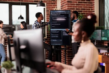 Afrika kökenli Amerikan sistem mühendisleri kaynak kodları analiz edip hata var mı diye bakıyorlar. Ajans ofisindeki büyük televizyon monitörlerini kontrol ediyorlar. Yazılım geliştiricileri kurumda siber güvenlik yenilikleri üzerinde çalışıyorlar..