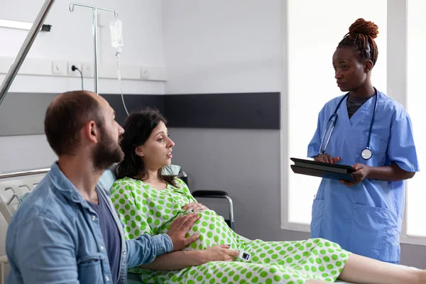 アフリカ系アメリカ人のアシスタントは妊娠中の女性と出産プロセスについて話し合い 病院病棟での手術のための母親を準備する 妊娠中の患者は婦人科医を待っているベッドに横たわっています — ストック写真
