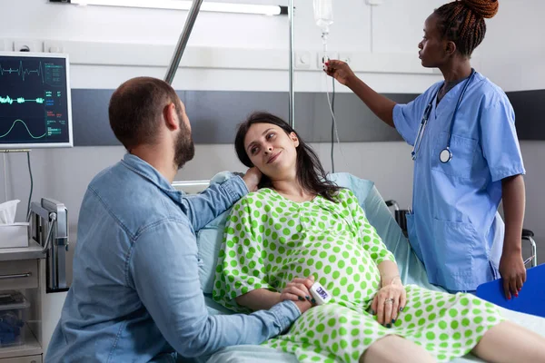 病棟で出産を待っている妊娠中のカップル アフリカ系アメリカ人看護師チェックIvドリップバッグ妊娠中の女性はベッドに横たわっていると夫は母親に慰め — ストック写真