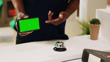 Müşteri telefonu yeşil ekranlı, lobideki resepsiyonistin izole edilmiş kromatonlu cep telefonu ekranını gösteriyor. Otel resepsiyonunda boş model şablon kullanan genç bir adam..