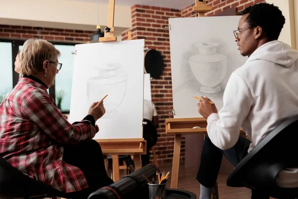 現代のスタジオでは 様々な世代の人々がキャンバスに絵を描き 創造的なアートレッスン中に話しています アフリカ系アメリカ人の学生の男に自然主義的な静物画の描き方を教える先輩 — ストック写真