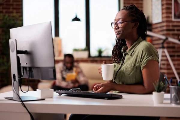 家里的程序员在客厅喝咖啡或喝茶时 用电脑轻松地在网上播放系列节目 非裔美国人自由职业者在Pc屏幕上花时间观看视频内容 — 图库照片
