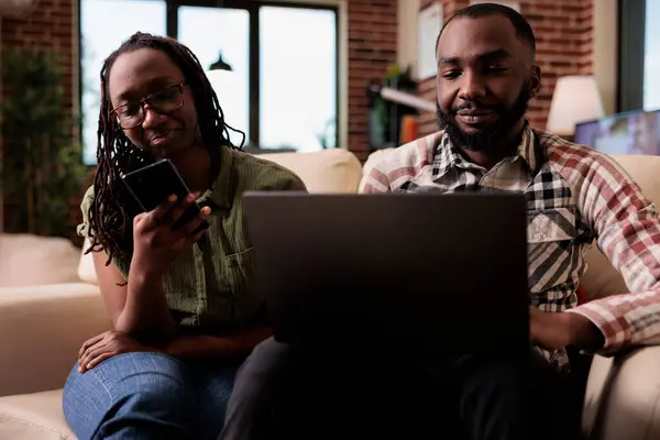 彼氏がソファに座ってノートパソコンで作業している間 アフリカ系アメリカ人女性はスマートフォンでソーシャルメディアを閲覧します 夫婦は自宅のリビングルームでデジタルデバイスを使用して一緒に自由な時間を過ごす — ストック写真