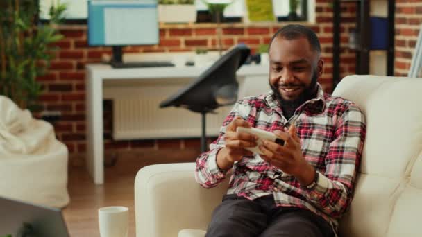 兴奋的非洲裔美国人在智能手机上玩电子游戏 在漫长的工作后放松了一天 在时尚的公寓客厅里打电话寻开心的激烈竞争的游戏玩家 — 图库视频影像