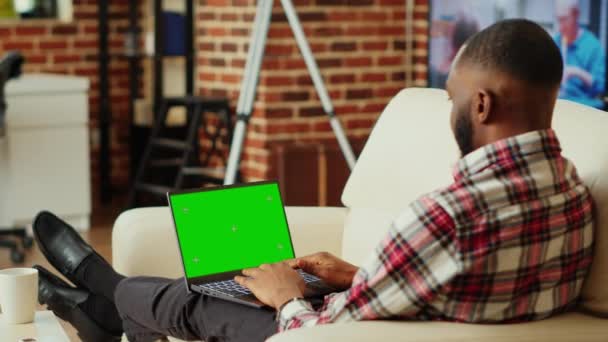 自宅で作業するリモートフリーランスに焦点を当て クロマキーにデータを入力すると ノートパソコンの緑の画面をモックします アフリカ系アメリカ人のテレワーカーが居心地の良いアパートのリビングルームで会社のタスクを解決 — ストック動画