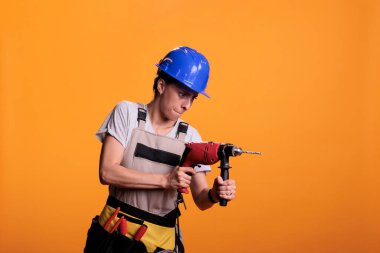Yeni iç yenileme projesinde matkap tabancası tutan kadın endüstriyel mühendis, elektrikli matkapla çivi tabancası kullanıyor. Stüdyoda üniforma giyen kadın, sarı arka planda çekim yapıyor..