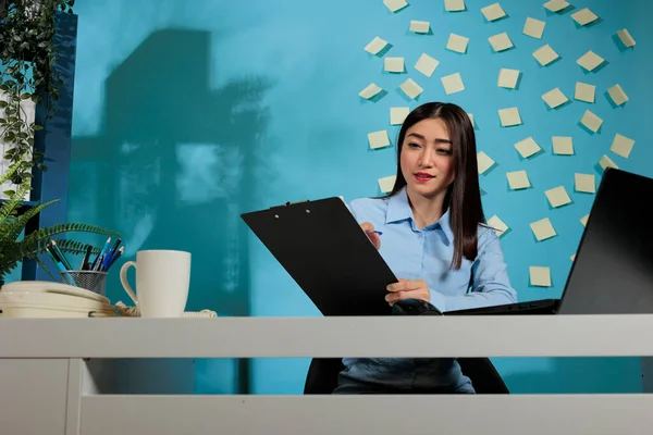 Corporate asian employee using laptop to do an online research and taking notes on clipboard. Businesswoman sitting at office desk concentrating on taking notes filling out form.