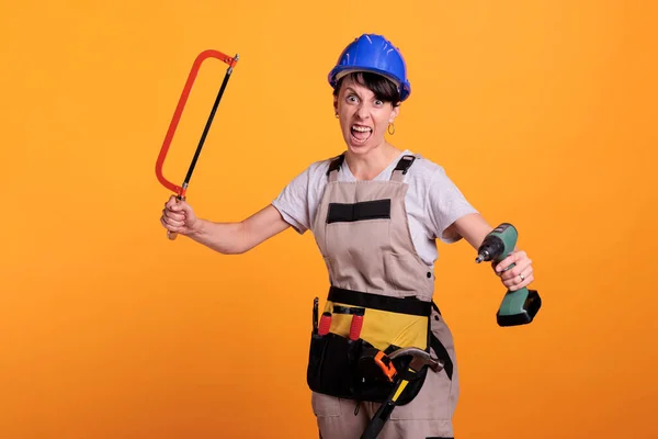 Angry violent woman builder with chainsaw and power drill acting crazy in studio, posing with building tools. Mad female worker being frustrated with manual saw and drilling gun.