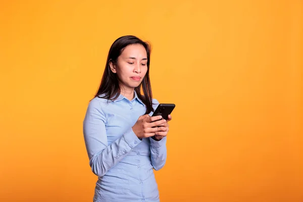 stock image Asian woman chatting with remote friend using smartphone, typing message during work break in studio over yellow background. Cheerful young adult browsing on social media, searching information