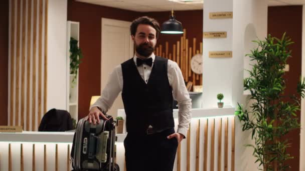 旅行リゾートラウンジで荷物トロリーで立って幸せな笑顔のベルボーイの肖像画 チェックイン受付に立っている陽気なエレガントなプロのホテルスタッフの従業員 — ストック動画