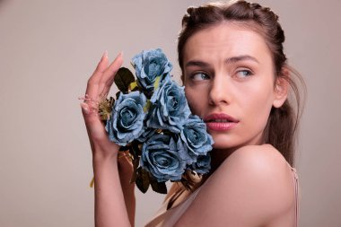 Zarif sarışın kadın elinde gülleri yüzüne yakın tutuyor ve yan tarafa bakıyor. Bej arka planda çiçek demeti ile poz veren narin genç ve güzel bir bayan.