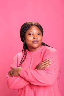Stüdyoda poz verirken güvenle gülümseyen neşeli bir kadının portresi çekimlerden zevk alıyor. Afro-Amerikan model, şık saç stili ve pembe tüylü kameraya bakıyor.