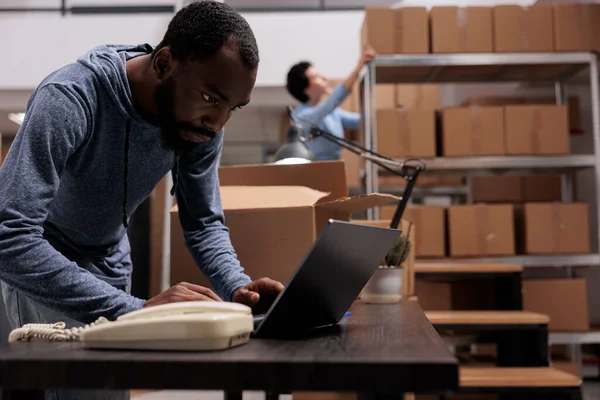 アフリカ系アメリカ人従業員は 顧客の注文の準備を開始する前に貨物在庫を見て 倉庫配送部門で働いています 労働者のラップトップコンピュータ上の出荷の詳細をチェック — ストック写真