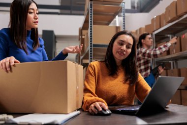 Depo iki Asyalı kadın işçi dizüstü bilgisayardan paket teslimat programı planlıyorlar. Depo çalışanları depolama odasındaki bilgisayarda mal yönetimini kontrol ediyor.