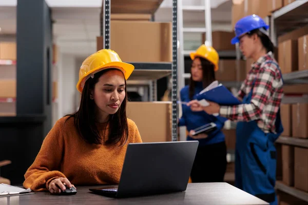 ラップトップ上の小包追跡システムを使用して郵便サービス倉庫労働者 若いアジアの倉庫の従業員は デスクで製品の物流や流通を管理する黄色のハード帽子を着用 — ストック写真