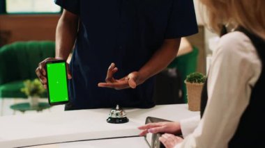 Otelin misafiri telefonda yeşil ekranı gösteriyor, akıllı telefondan izole edilmiş görüntülere bakıyor. Resepsiyonda boş kromakey şablonu olan Afrika kökenli Amerikalı adam tutma cihazı..