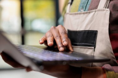 Depo çalışanı dizüstü bilgisayardan müşteri siparişlerini kontrol ediyor, depo odasında nakliye için malzeme hazırlıyor. Afro-amerikan çalışan depoda mal lojistiği analizi yapıyor. Kapat.