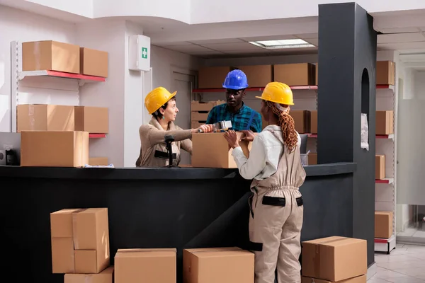 倉庫労働者チームは郵便局の収納受付で粘着テープディスペンサーで段ボール箱を閉じます 配送サービスマネージャーは一緒に接着剤で小包を梱包 — ストック写真