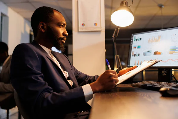 執行役員は コンピュータ上のビジネスデータと比較して企業統計で論文を分析します アフリカ系アメリカ人起業家 スタートアップオフィスのマーケティング戦略で残業 — ストック写真