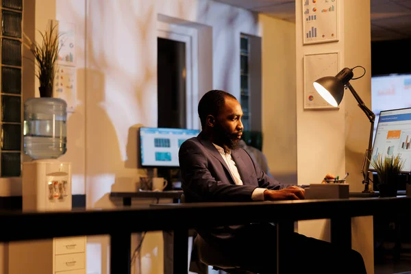 非裔美国商人站在办公桌前分析计算机上的财务图表 深夜在创业办公室为公司战略工作 执行经理规划投资计划 — 图库照片