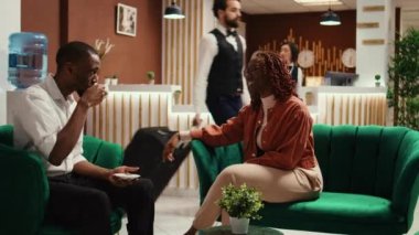 Afrikalı Amerikalı çift otel lobisinde bir fincan kahvenin tadını çıkarırken rezervasyon onayını bekliyor. Neşeli, rahat turistler modern lüks dinlenme odasında rezervasyon yaptırmaya hazır.