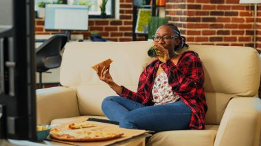 Happy woman eating pizza from delivery at home, enjoying movie on television with beer bottles and fast food takeaway. Modern person having fun binge watching show on tv, takeout.